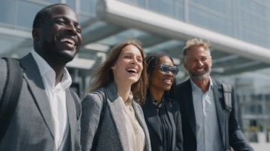 Vier berufstätige Frauen und Männer unterschiedlicher Herkunft lachen gemeinsam vor einem modernen Gebäude. Sie tragen Businesskleidung und haben Reisegepäck dabei.