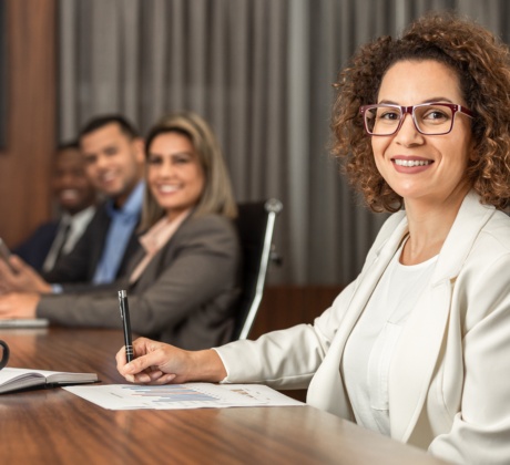 Eine Geschäftsfrau im cremefarbenen Blazer und viereckiger Brille sitzt an einem Versammlungstisch. Sie trägt braune Locken und lächelt selbstbewusst in die Kamera. Vor ihr auf dem Tisch liegt ein Blatt Papier, ein geöffnetes Notizbuch und eine schwarze Kaffeetasse. Im Hintergrund sind weitere Kollegen zu erkennen, die ebenfalls in Richtung der Kamera lächeln.