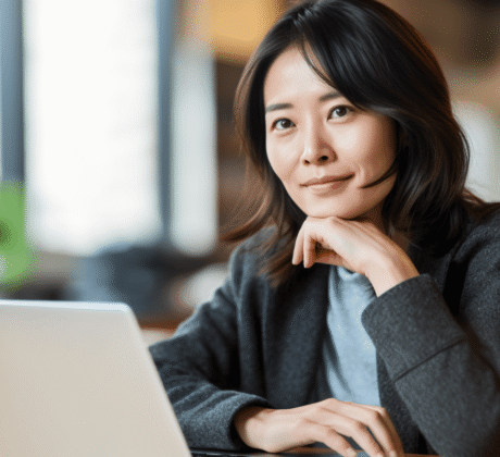 Asia-Deutsche Frau sitzt nachdenklich und zufrieden am Schreibtisch. Sie stützt ihren Kopf mit der Hand und schaut auf ihren Laptop.