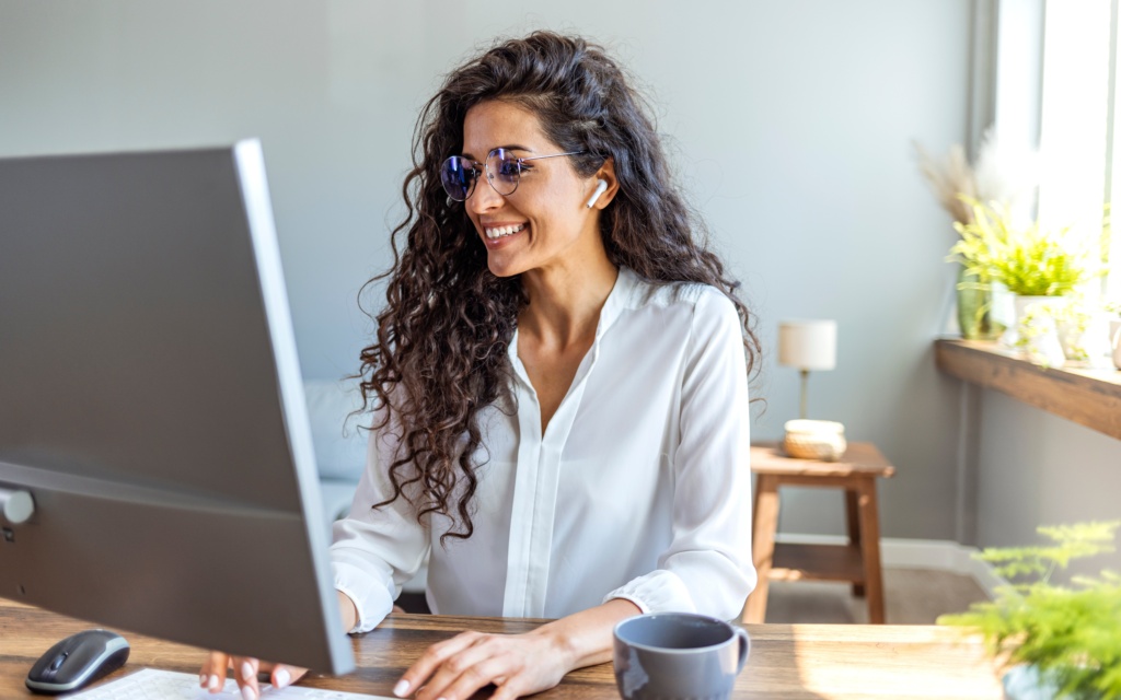 Junge Frau sitzt lächelnd am Computer und tippt in die Tastatur. Vor ihr steht eine graue Tasse. Zwei Zimmerpflanzen sind zu sehen. Das Fenster ist daneben und die Sonne scheint ins Zimmer. Die Wand ist in Pastellfarben gestrichen. Die Junge Frau trögt die gelockten Haare offen, zu einer Seite fallen sie über die Schulter. Sie trägt eine runde große Brille und eine weiße Bluse. Hinter ihr nutzt sie einen kleinen Holzhocker als Abstellfläche für ihre Lampe.