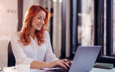 Eine Frau mit roten Haare sitzt vor dem Laptop und tippt fröhlich. Sie trägt eine weiße Bluse