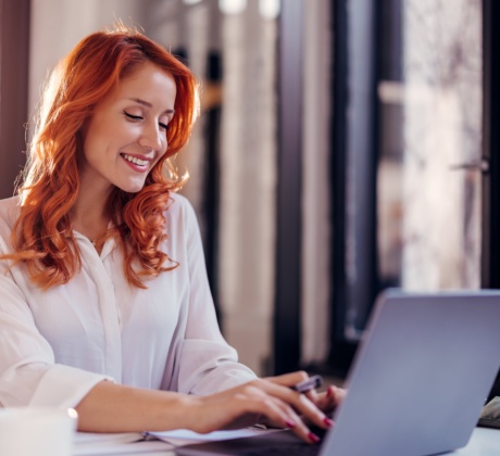 Eine Frau mit roten Haare sitzt vor dem Laptop und tippt fröhlich. Sie trägt eine weiße Bluse