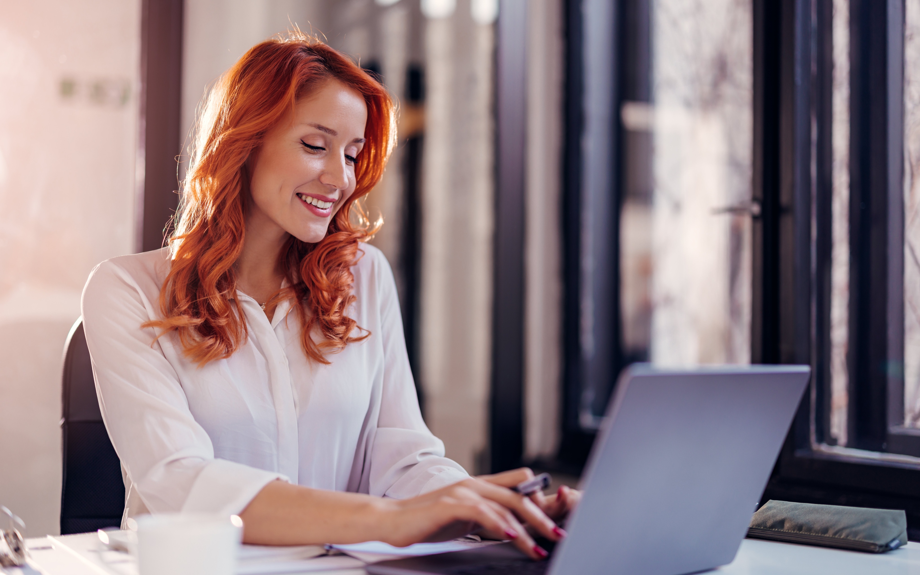 Eine Frau mit roten Haare sitzt vor dem Laptop und tippt fröhlich. Sie trägt eine weiße Bluse
