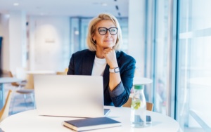 Eine Businessfrau in dunkelblauen Blazer und weißem Shirt sitzt in einem modernen Gebäude an einem runden weißen Tisch neben einer Fensterfront. Sie trägt ihre blonden Haare in einer Bobfrisur und trägt eine schwarze Brille. Ihr Blick geht nach draußen und sie hat ihren Kopf auf ihre Hand gestützt. An ihrem Handgelenk trägt sie eine weiße Armbanduhr. Vor ihr auf dem Tisch steht ein aufgeklapptes Notebook, ein dunkelblaues Notizbuch und eine Wasserflasche.