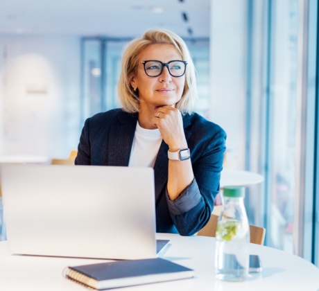 Eine Businessfrau in dunkelblauen Blazer und weißem Shirt sitzt in einem modernen Gebäude an einem runden weißen Tisch neben einer Fensterfront. Sie trägt ihre blonden Haare in einer Bobfrisur und trägt eine schwarze Brille. Ihr Blick geht nach draußen und sie hat ihren Kopf auf ihre Hand gestützt. An ihrem Handgelenk trägt sie eine weiße Armbanduhr. Vor ihr auf dem Tisch steht ein aufgeklapptes Notebook, ein dunkelblaues Notizbuch und eine Wasserflasche.