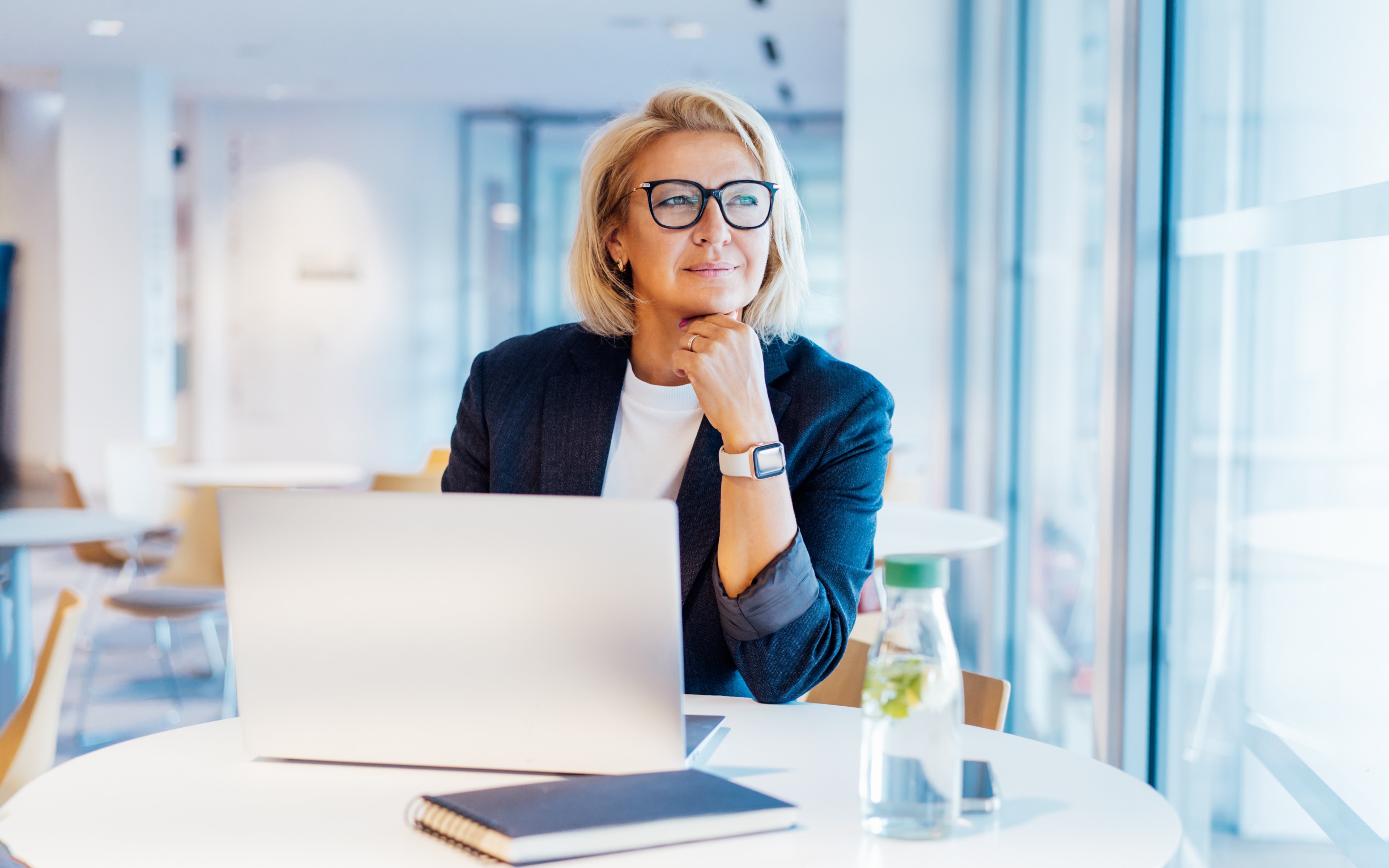 Eine Businessfrau in dunkelblauen Blazer und weißem Shirt sitzt in einem modernen Gebäude an einem runden weißen Tisch neben einer Fensterfront. Sie trägt ihre blonden Haare in einer Bobfrisur und trägt eine schwarze Brille. Ihr Blick geht nach draußen und sie hat ihren Kopf auf ihre Hand gestützt. An ihrem Handgelenk trägt sie eine weiße Armbanduhr. Vor ihr auf dem Tisch steht ein aufgeklapptes Notebook, ein dunkelblaues Notizbuch und eine Wasserflasche.