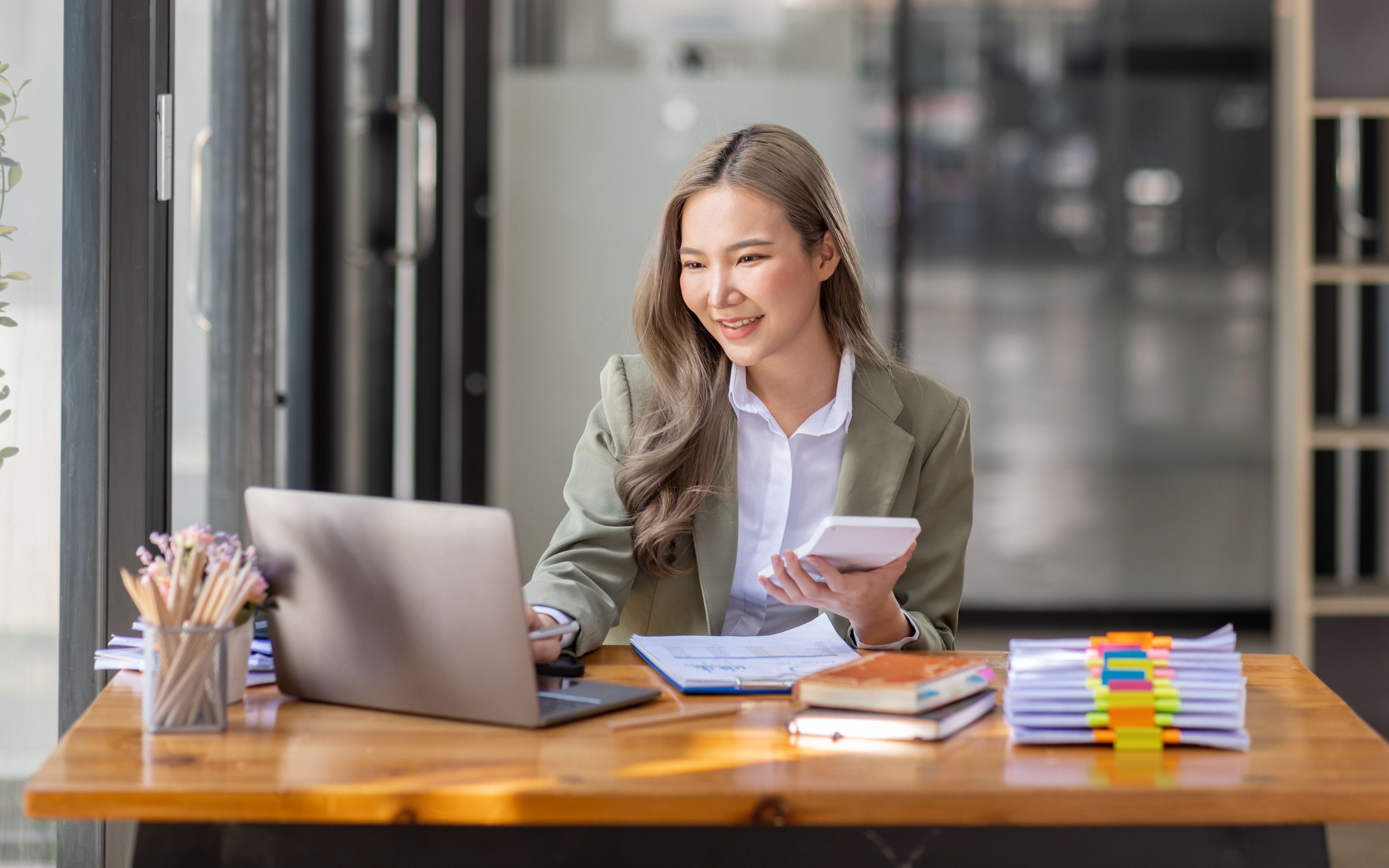 Eine Frau in einem jadegrünem Blazer und weißer Bluse sitzt an einem Holzschreibtisch. Sie tippt lächelnd mit einer Hand auf einem Laptop, in der anderen hält sie einen Taschenrechner. Auf dem Schreibtisch mehrere Unterlagen, Dokumente, ein Stifthalter mit vielen Bleistiften. Im Hintergrund steht ein minimalistisches Holzregal und eine Glasfront.