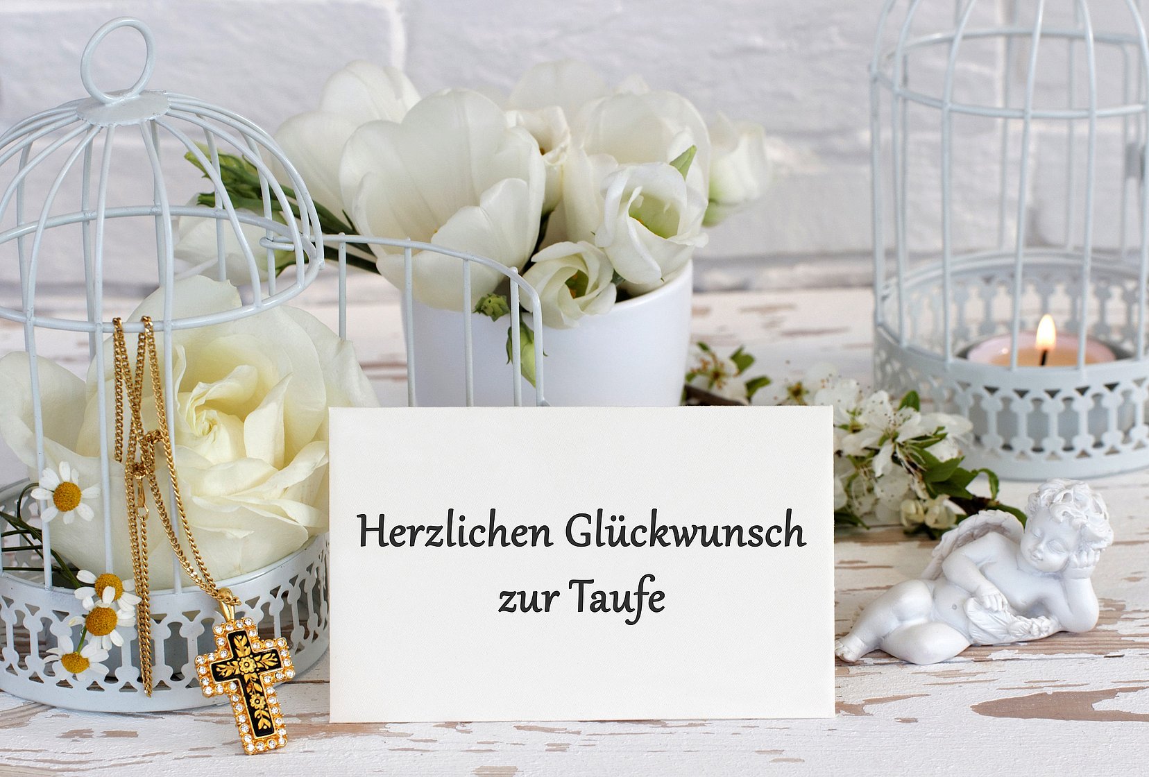 Im Mittelpunkt des Bildes steht eine weiße Vase  mit einem Strauß weißer Rosen. Rechts davon steht eine weiße Laterne mit einem brennenden Teelicht. Links neben der Vase steht eine weitere weiße Laterne mit einer weißen Rose und einer goldenen Kette mit einem goldenen Kreuz als Anhänger. In der Mitte vor der weißen Vase steht eine Karte mit der Aufschrift