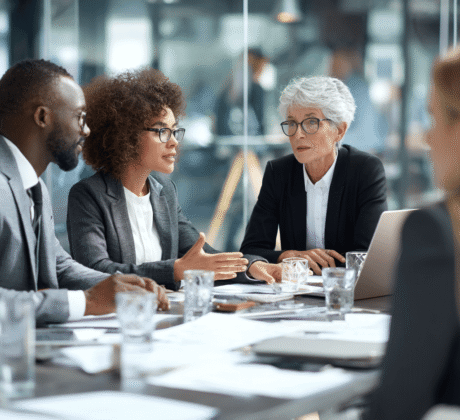 Vier Personen in Businesskleidung sitzen an einem Konferenztisch mit Dokumenten und Laptop; eine Frau spricht gestikulierend, die anderen hören aufmerksam zu.
