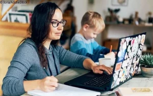 Eine Frau sitzt an einem Tisch mit Ihrem Laptop und arbeitet, während Ihr Sohn neben Ihr sitzt und was malt. Sie trägt einen graublauen Pulli und eine schwarze Brille.