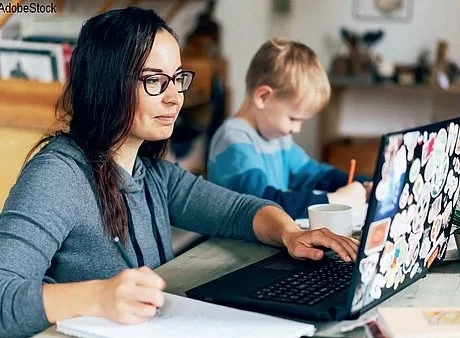 Eine Frau sitzt an einem Tisch mit Ihrem Laptop und arbeitet, während Ihr Sohn neben Ihr sitzt und was malt. Sie trägt einen graublauen Pulli und eine schwarze Brille.