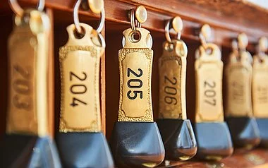 Nahaufnahme von mehreren schweren, gold-schwarzen Hotelschlüsseln, die ordentlich an Haken hängen. Die Schlüsselanhänger sind mit Zimmernummern wie 204, 205, 206 und 207 beschriftet.