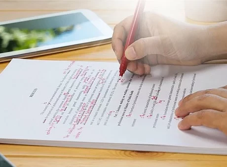 Ein Block liegt auf dem Tisch und eine Person korrigiert den Text auf diesen mit einem roten Stift. Zu sehen sind nur die Hände der Person.