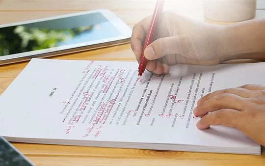 Ein Block liegt auf dem Tisch und eine Person korrigiert den Text auf diesen mit einem roten Stift. Zu sehen sind nur die Hände der Person.