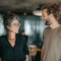 Kollegin mit Brille und hochgesteckten Haaren spricht freundlich mit jüngerem Kollegen mit Man-Bun im modernen Büro – Symbol für gelungene Bürokommunikation.