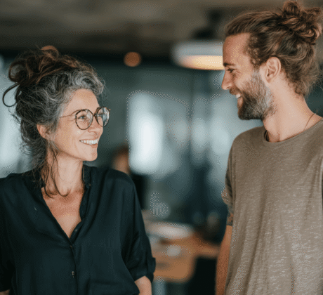 Kollegin mit Brille und hochgesteckten Haaren spricht freundlich mit jüngerem Kollegen mit Man-Bun im modernen Büro – Symbol für gelungene Bürokommunikation.