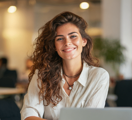 Fröhliche Frau mit langen lockigen Haaren und dunklen Augen arbeitet am Laptop in einem hellen, modernen Büro, freundliche und professionelle Atmosphäre.