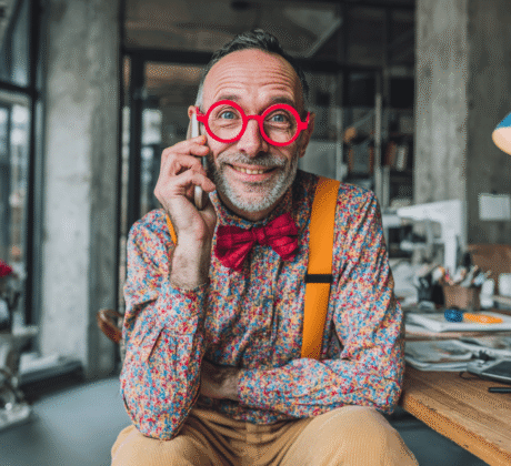 Schlanker Mann Anfang 40 mit Halbglatze, Bart, bunter Kleidung, Fliege, Cordhose, Hosenträgern und roter Hornbrille telefoniert lachend im modernen Industrial-Büro