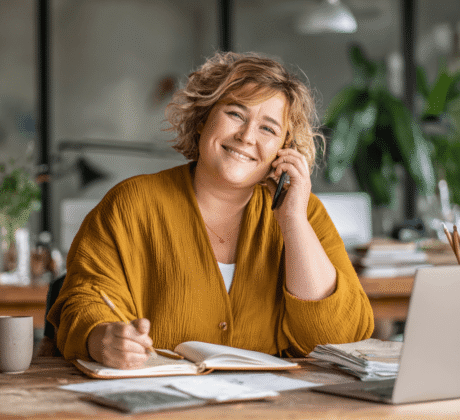 Plus-Size Frau im Büro telefoniert und schreibt Notizen – professionelle Korrespondenz am Schreibtisch.