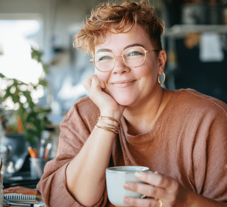 Sympathische Frau mit goldener Brille sitzt am Schreibtisch und trinkt Tee oder Kaffee – Symbol für gesundes Trinken am Arbeitsplatz
