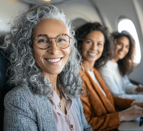 Drei selbstbewusste Frauen Anfang 50 sitzen während ihrer Geschäftsreise nebeneinander im Flugzeug; sie wirken professionell und entspannt, eine ideale Darstellung moderner Dienstreisen. Im Kontext steht eine praktische Checkliste für erfolgreiches Business-Travel-Management.