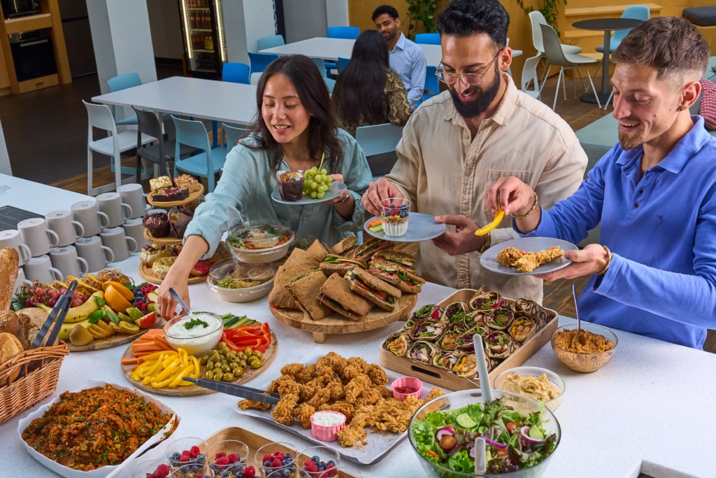 Drei Kolleginnen und Kollegen stehen an einem reich gefüllten Buffet mit Sandwiches, Salaten, Hähnchen, Dips und Früchten. Sie greifen nach Speisen und füllen sich ihre Teller.