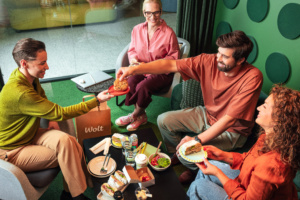 Vier Personen sitzen auf Sesseln um einen kleinen Tisch, auf dem Sandwiches, eine Bowl und Getränke stehen. Eine Person reicht einer anderen einen Cookie auf einem kleinen Teller.