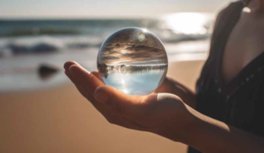 Eine Hand hält eine Glaskugel am Strand, in der sich der Himmel und das Meer auf dem Kopf stehend spiegeln.
