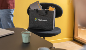 Eine graue Desk Sharing Bag mit Tragegriffen und einem Platzhalter-Logo („YOUR LOGO“) steht auf einem schwarzen Bürostuhl.